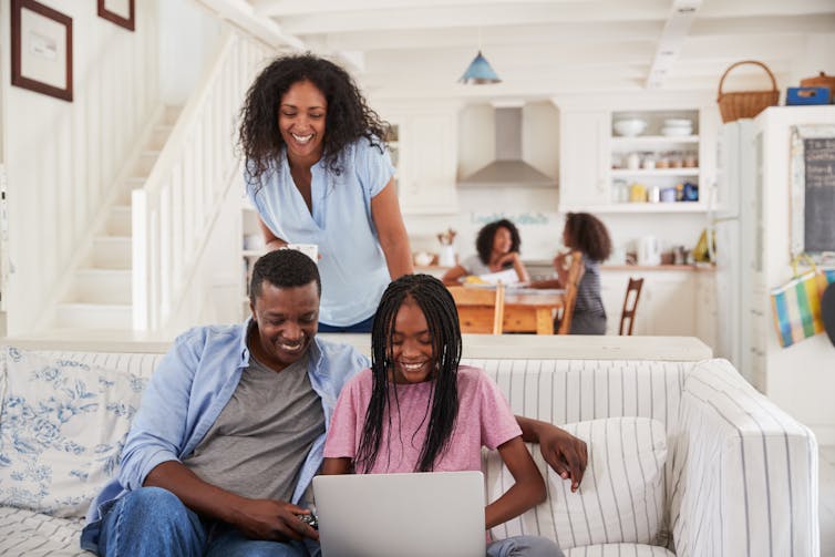 A man and teen girl sit on a couch using a laptop computer, a woman stands behind them.