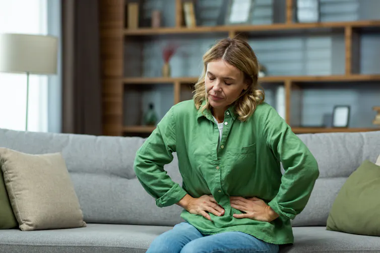 Woman leaning forward on couch with eyes closed in discomfort, hands pressing against stomach