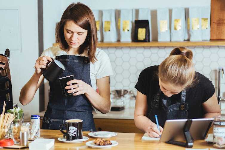 Two young women work behind a cafe counter. One pours milk into a cup, another writes on a pad.