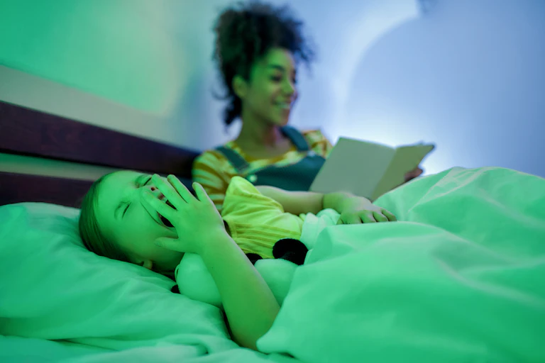 A child in bed yawning with a babysitter in the background, green and blue lighting.