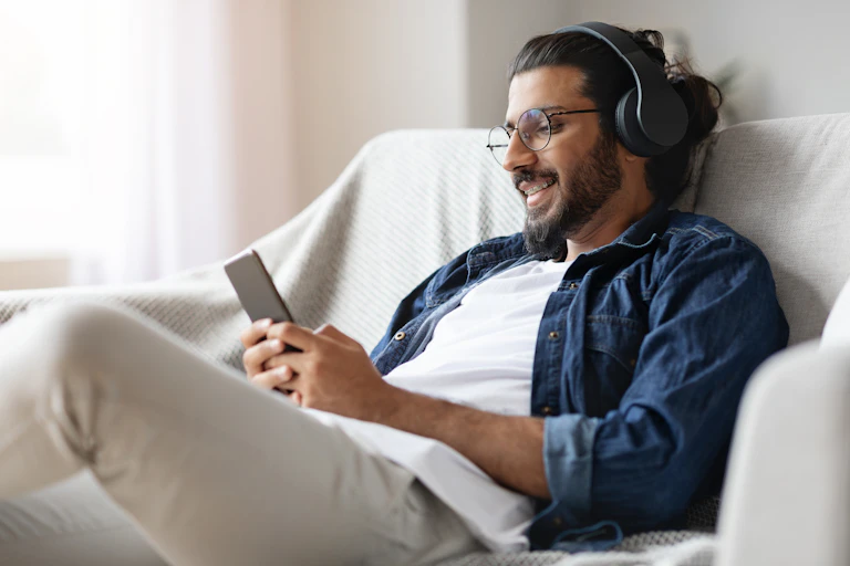 A young man sitting on a couch, smiling and listening to headphones while looking at his mobile phone