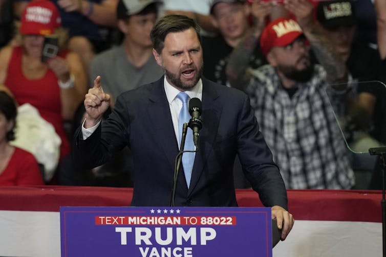 A middle-aged white man in a suit gestures while speaking to a crowd from behind a podium that says 'Trump Vance' on the front