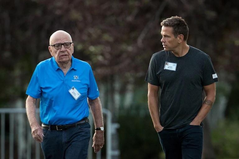 Two casually dressed white men -- one elderly, one middle-aged -- converse while on a walk.