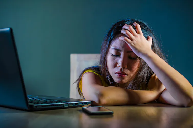 A girl with a distressed look on her face rests her forehead against her hand in front of an open laptop