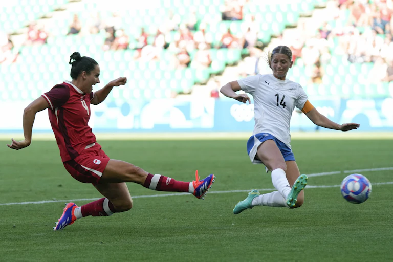 A woman in a red jersey kicks a ball while another in white attempts to deflect it