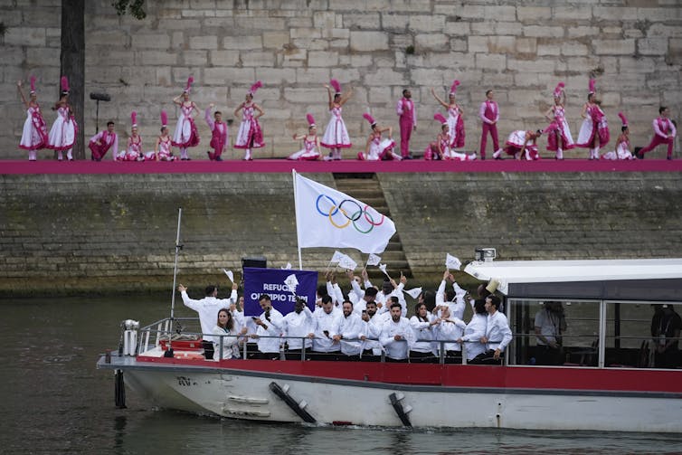 A boat with athletes in white, and dancers in pink in the background.