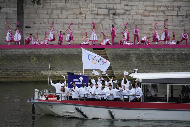 A boat with athletes in white, and dancers in pink in the background.