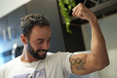 A man with dark, curly hair flexes his bicep, which has a tattoo of the Olympic rings on it