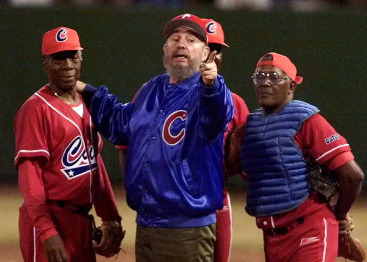 An older man with a full beard and red ball cap grasps the shoulder of a man in a red baseball uniform while two other players stand behind them