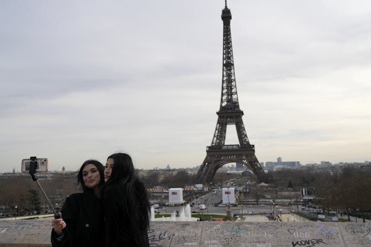 Two women take a photo using a selfie stick, the eiffel tower is seen in the background.