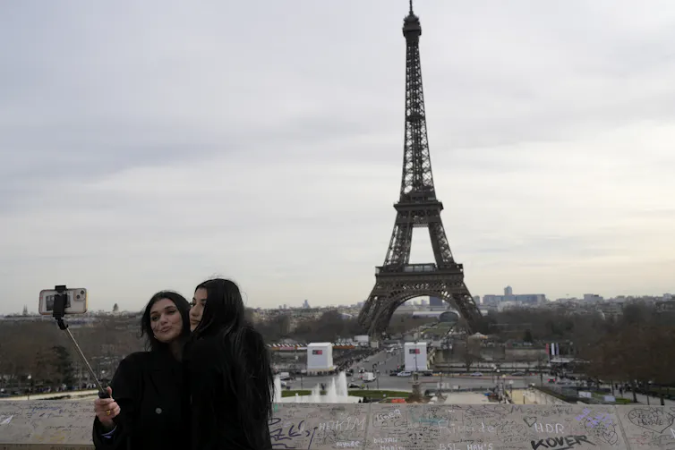 Two women take a photo using a selfie stick, the eiffel tower is seen in the background.
