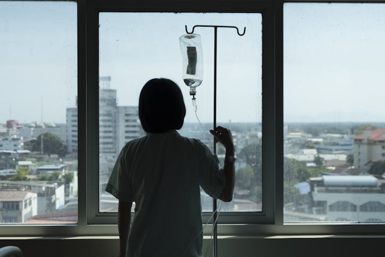 A woman connected to an IV drip looks out a hospital window.