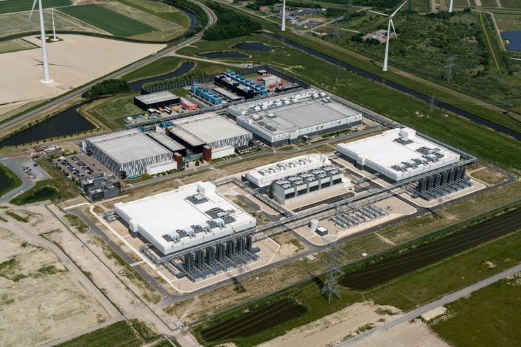 Aerial view of several white rectangular buildings surrounded by wind turbines and green fields.