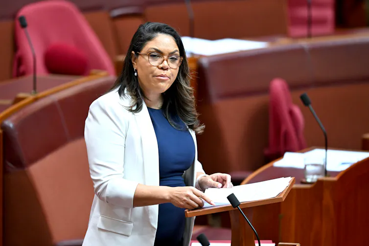 A woman in a white blazer stands in parliament and speaks
