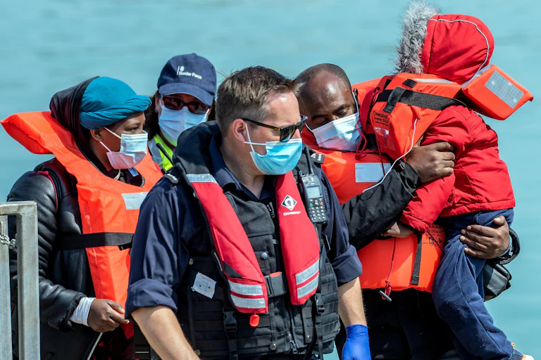 A Border Force official walking in front of three migrants, a woman, man and a child, all wearing life vests and face masks