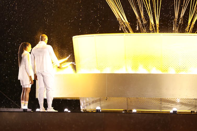 Two athletes in white light the Olympic cauldron.