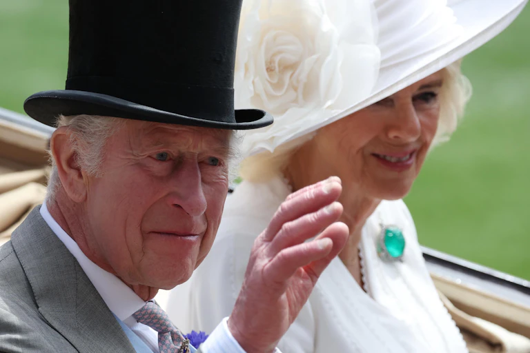 King Charles wearing a top hat sits beside Queen Camilla, wearing an elaborate white hat. They are sitting in an open top carriage and smiling, the king is waving