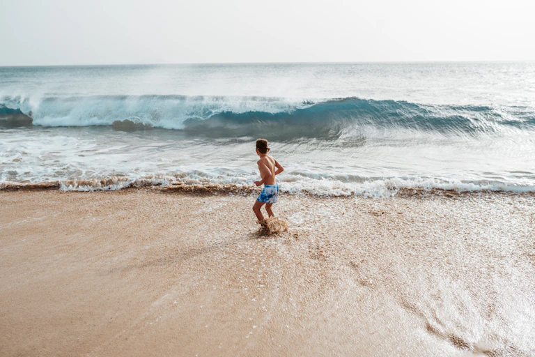 Un enfant hésite à se jeter dans les vagues, sur une plage face à la mer.