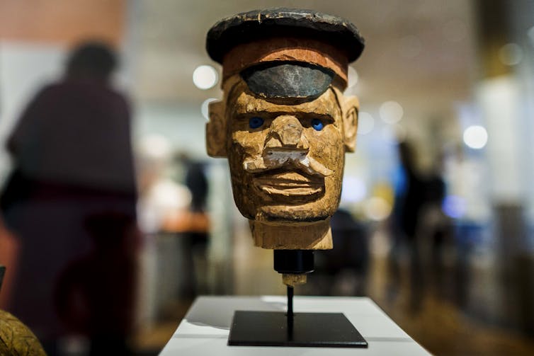 Une figure sculptée d'un homme portant une casquette militaire, avec des yeux bleus perçants, une grosse moustache et un sourire menaçant