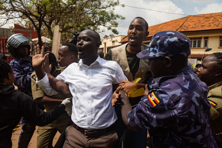 People in camouflage uniform and a brown uniform hold the arms of a man in civilian clothes