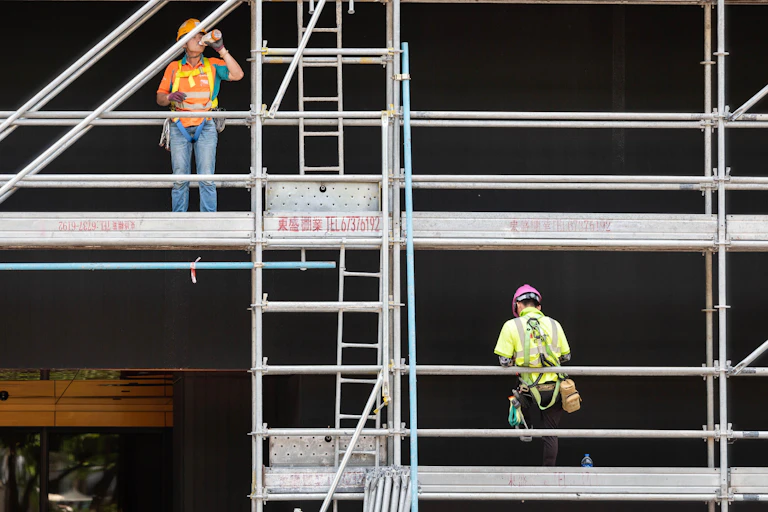 two construction workers in high vis and hard hats on scaffold, one drinking a drink
