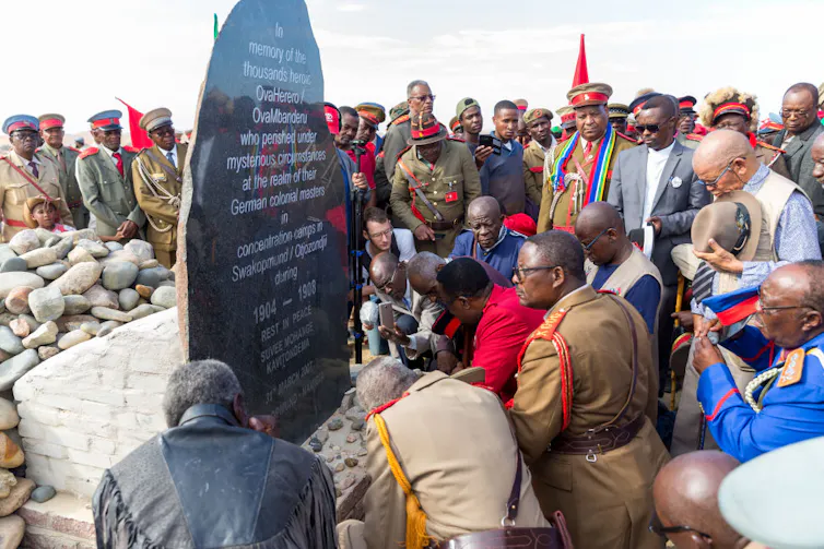 Men, some in military uniforms, gather and kneel solemnly at a gravestone marking an account of genocide.
