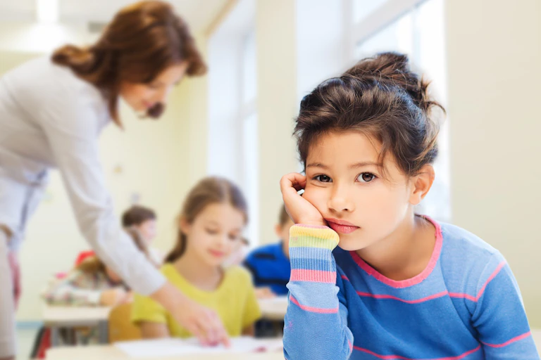 Girl in foreground looking sad while teacher helps child in background