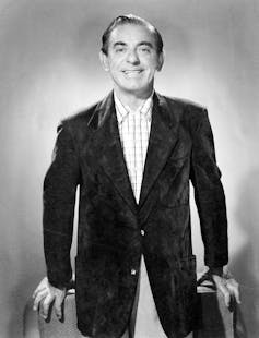 A black and white portrait of a smiling man in a checked shirt and black jacket smiling against a light-colored backdrop.
