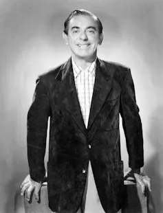 A black and white portrait of a smiling man in a checked shirt and black jacket smiling against a light-colored backdrop.