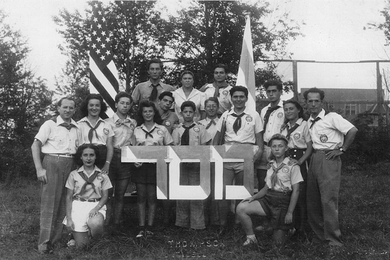 Three rows of young people in short-sleeve button-up shirts and scarves pose with two flags outside.