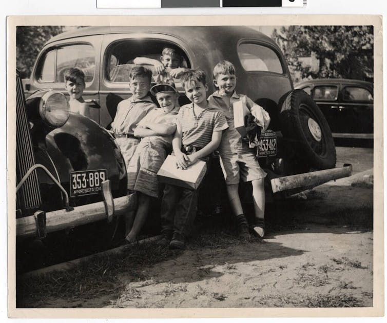 Half a dozen young boys stand in and around small cars in a black and white photograph.