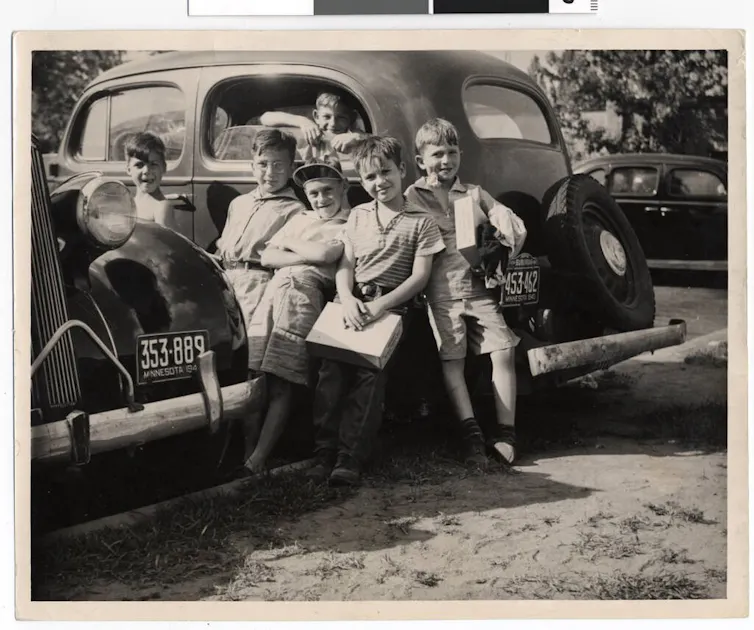 Half a dozen young boys stand in and around small cars in a black and white photograph.