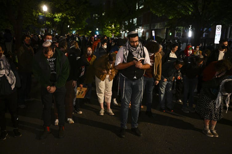 Rows of students stand in prayer on pavement outside at night.