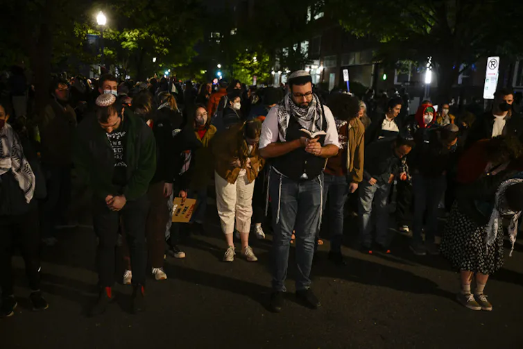 Rows of students stand in prayer on pavement outside at night.