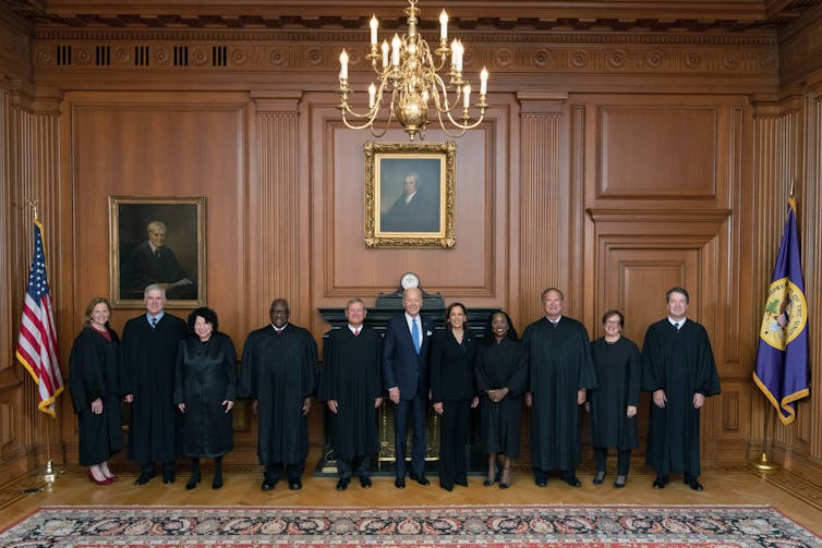 A group of people, most of whom are wearing black robes, stand shoulder to shoulder in a formal looking room with a carpet and wood panels on the walls.
