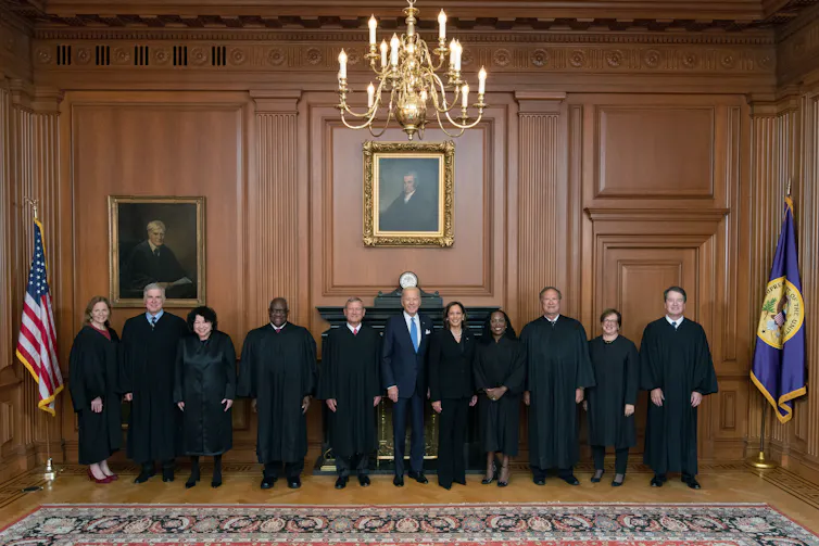 A group of people, most of whom are wearing black robes, stand shoulder to shoulder in a formal looking room with a carpet and wood panels on the walls.