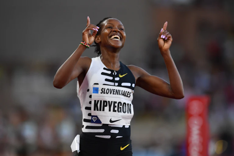 Une femme africaine vêtue d'une veste d'athlétisme se tient les bras levés, l'index pointé vers le haut en signe de victoire.