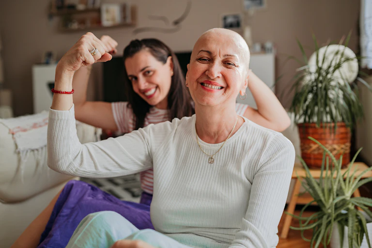Two women, one of whom is going through cancer treatment, raise their fists in a determined gesture.