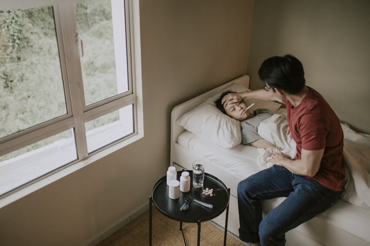 A sick boy lies in bed as his father takes his temperature.