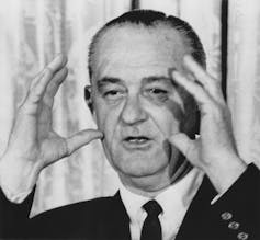 A man speaking with his hands at the side of his head.