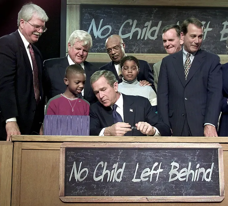 A man seated at a desk surrounded by five other men in suits and two young boys in regular clothes.