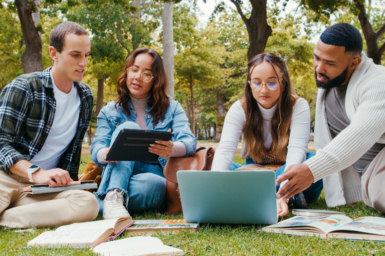 Four adult students sit on the grass with books and a laptop.
