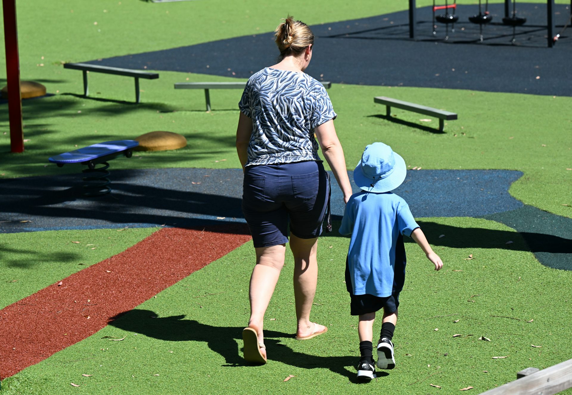 A woman leads a small child in school uniform across a playground.