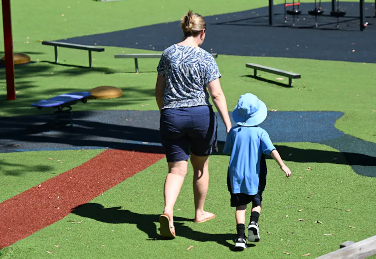 A woman leads a small child in school uniform across a playground.