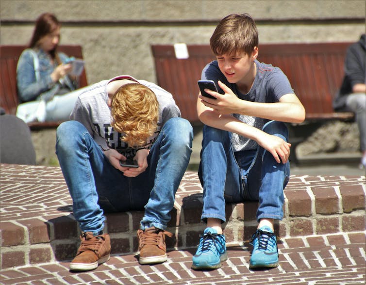 Two teenage boys sitting on the pavement using smartphones.