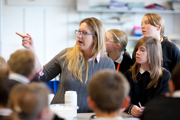 A woman points to the left. She is surrounded by children in uniform in a classroom.