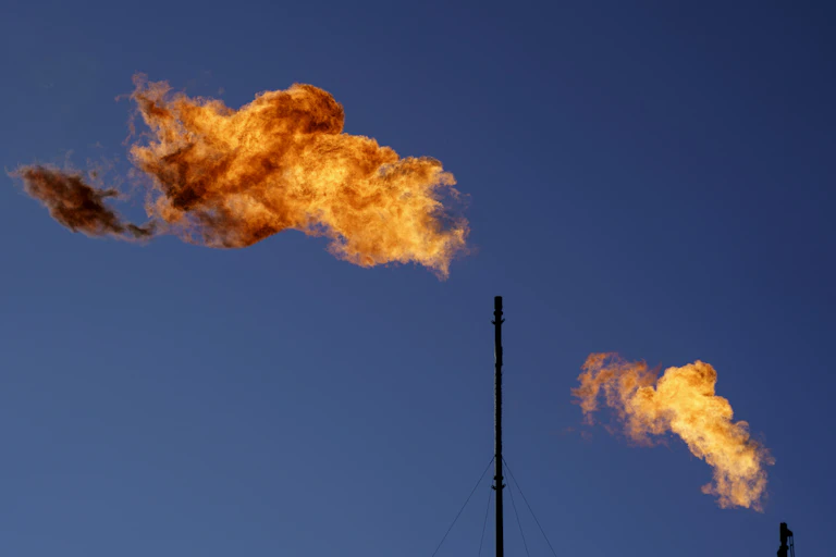 Flares burn off methane and other hydrocarbons at an oil and gas facility, two orange flames contrast against a blue sky