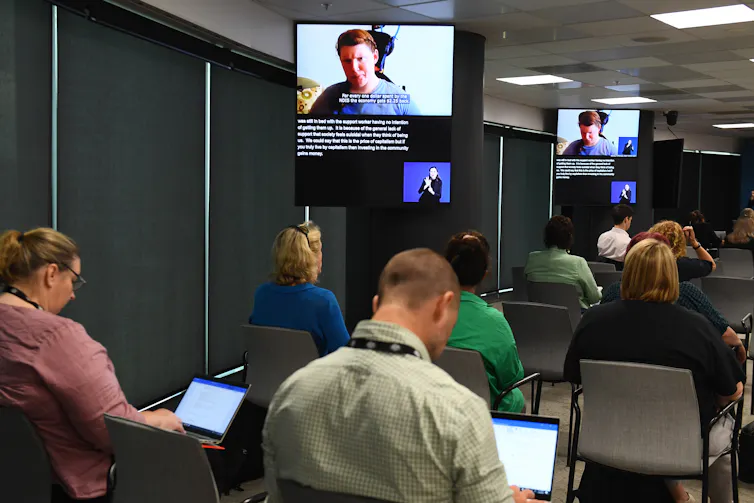 people sit in official room taking notes on computers while a woman appears via video link onscreen
