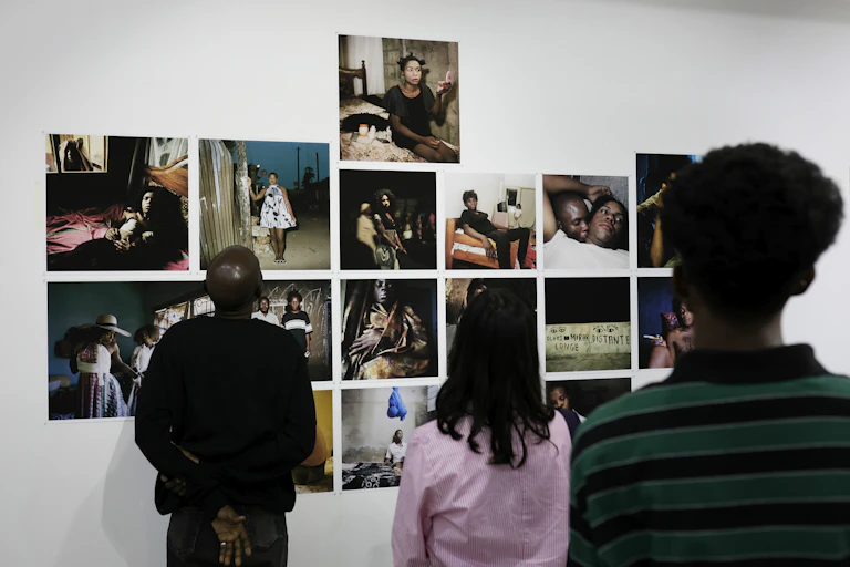 The backs of three people who are looking at a wall that's displaying multiple unframed photographs, mostly various styles of portraits of Africans.