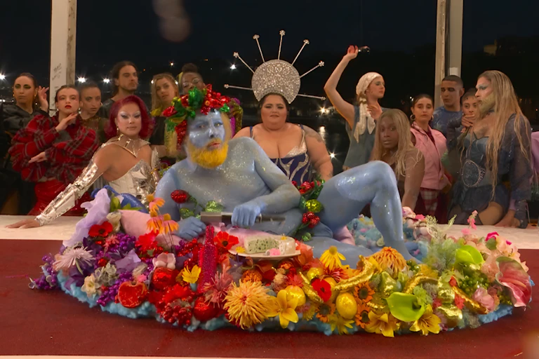 Un homme peint en bleu au front ceint d'une couronne de fleurs allongé sur une table couverte de fleurs ; derrière la table, une dizaine de personnes habillées de façon extravagante